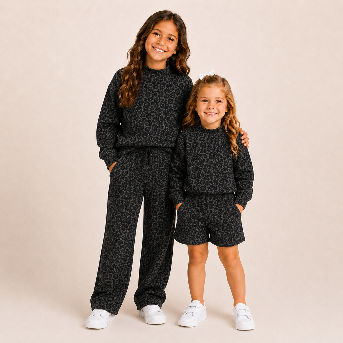 Two young girls modeling matching black and gray leopard-print sweatshirt and pants sets, standing against a solid beige studio background.