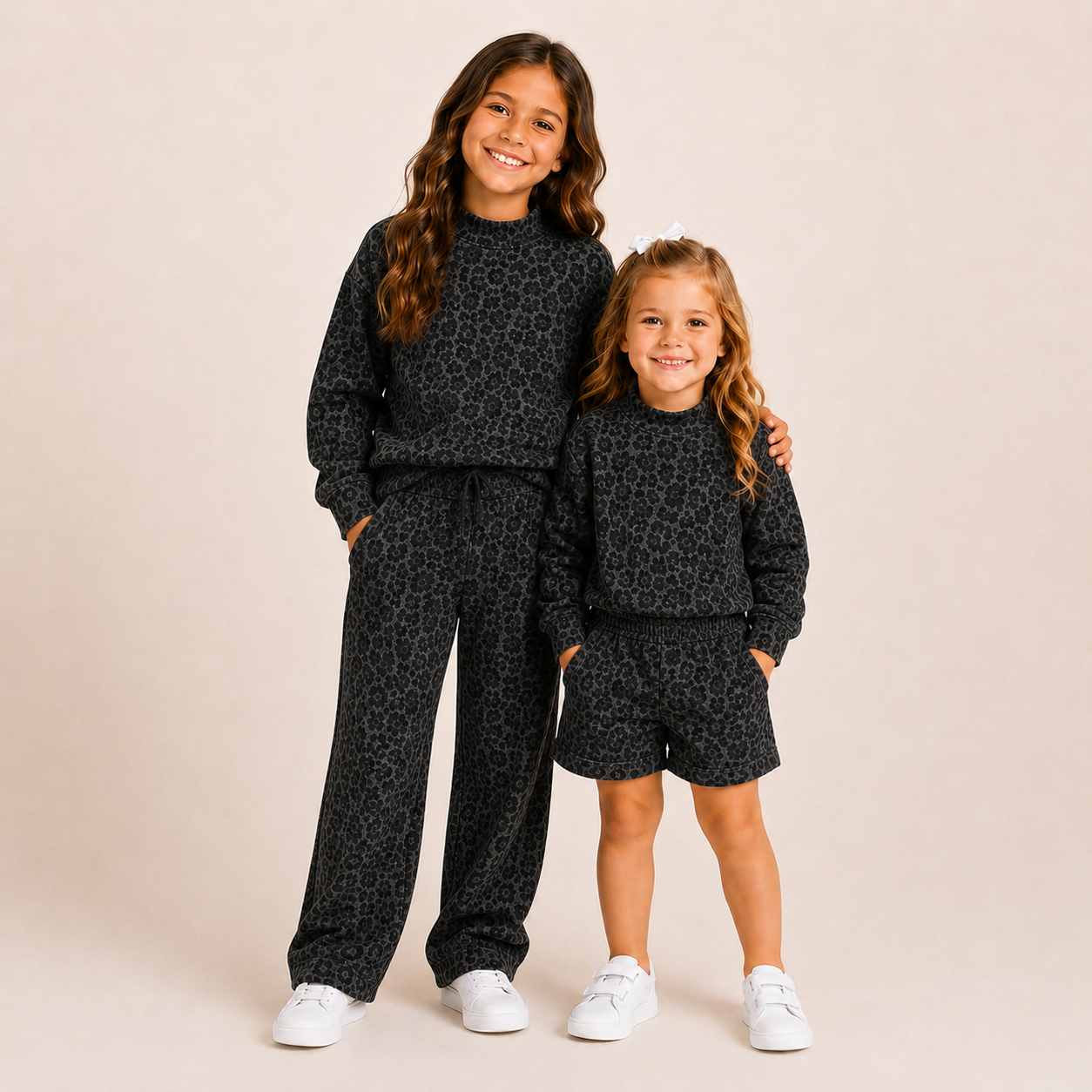Two young girls modeling matching black and gray leopard-print sweatshirt and pants sets, standing against a solid beige studio background.