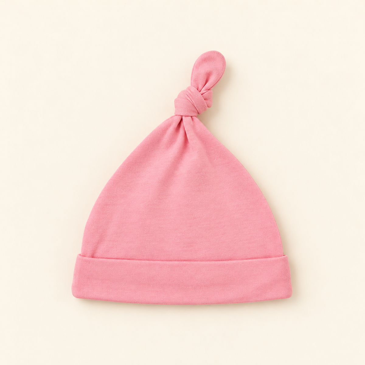A light pink knit baby beanie hat with a knotted top and folded cuff, displayed flat against a warm cream background.