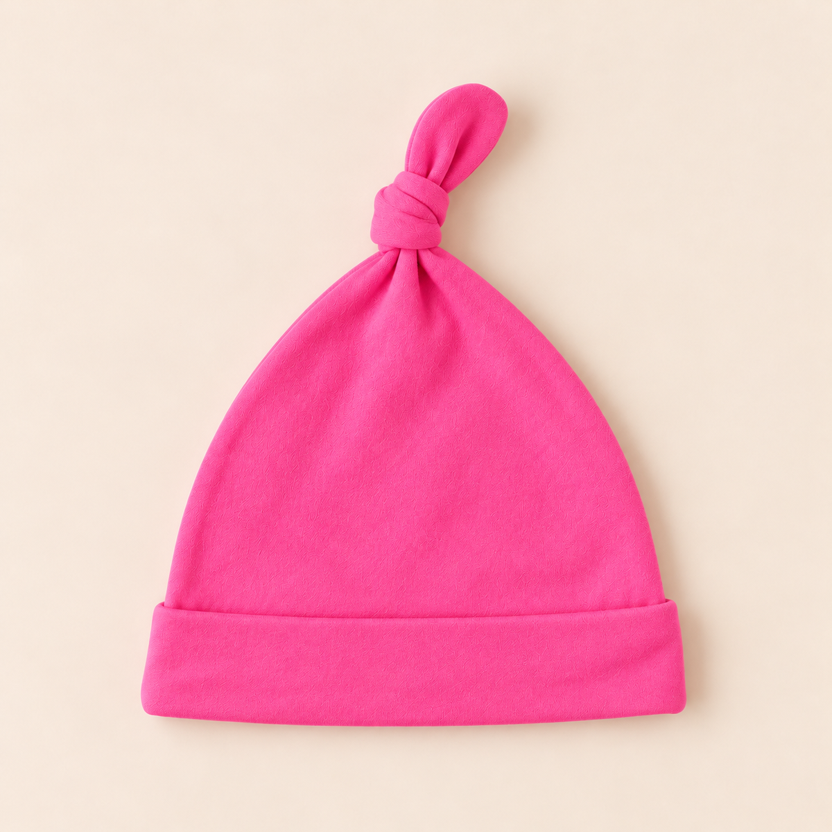 A bright pink knit baby beanie hat with a folded cuff and a knotted top, shown flat on a light beige background.