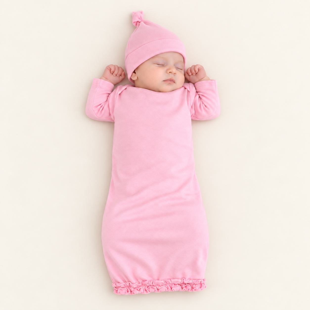 A sleeping newborn baby wrapped in a soft pink long-sleeve swaddle blanket with a matching pink beanie hat, lying on a warm off-white background.