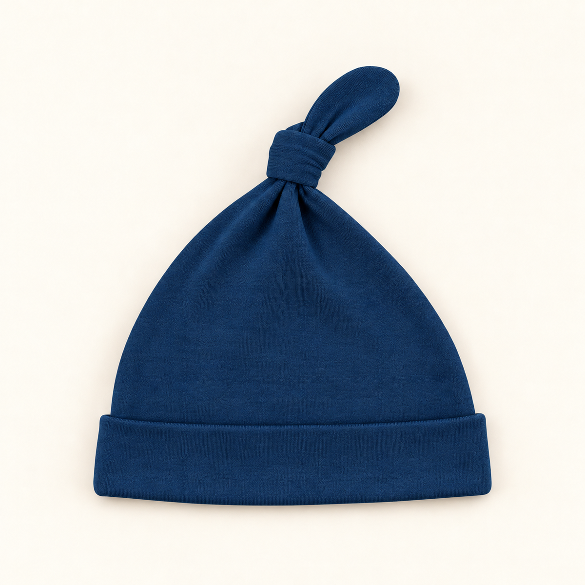 A dark blue knit baby beanie hat with a knotted top and folded cuff, photographed flat on a light cream background.
