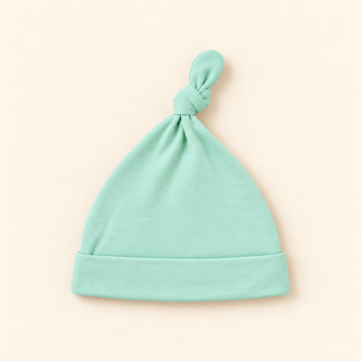 A mint green baby beanie hat with a knotted top and folded cuff, displayed flat on a light beige background.