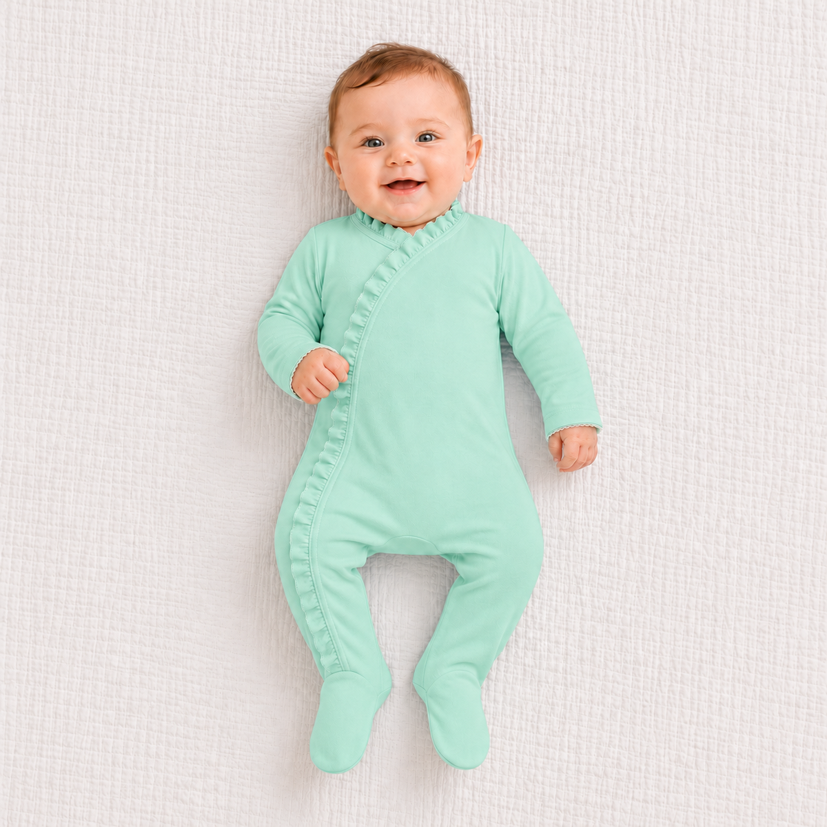 A baby lying on a textured white blanket wearing a mint green footed sleeper with a ruffled collar and side zipper.
