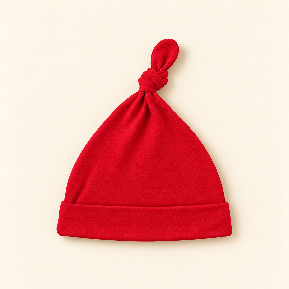 A bright red knit baby hat with a folded cuff and a knotted top, photographed flat on a warm cream background.